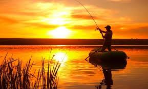 Fisherman casting rod at sunset on calm lake, golden sky reflecting on water, reeds in foreground