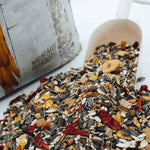 Natural bird seed mix with sunflower seeds, dried fruits, and grains spilling from a white scoop near a nature-themed bag