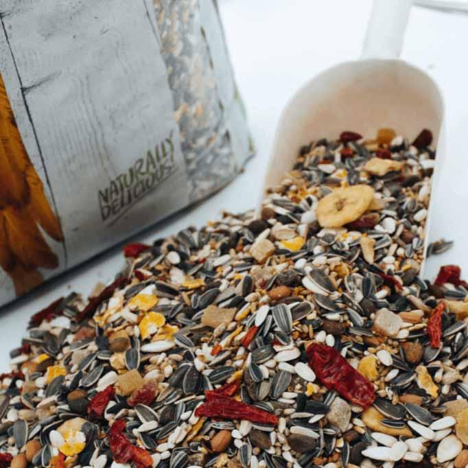 Natural bird seed mix with sunflower seeds, dried fruits, and grains spilling from a white scoop near a nature-themed bag
