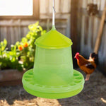 Green plastic hanging poultry feeder with chicken blurred in coop background